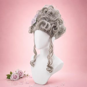 May include: A grey wig with elaborate curls and a small lavender flower accent, displayed on a white mannequin head. The wig features long, flowing curls and is set against a soft pink background. A few pink rose blossoms are at the base.