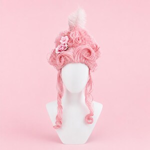 May include: A pink wig in a bouffant style, decorated with a white feather and two pink fabric flowers. The wig is displayed on a white mannequin head against a pink background. The wig has long, curled side pieces.
