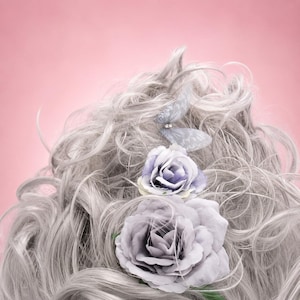 May include: A close-up shot of grey, wavy hair decorated with two lavender roses and a butterfly hair clip. The background is a solid pink. The hair has a soft, textured appearance, and the flowers add a touch of elegance.