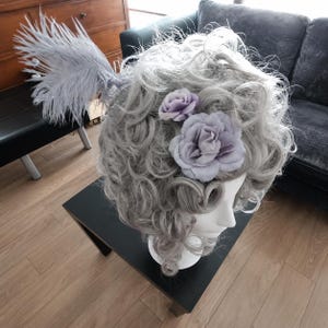 May include: A grey wig with large curls, decorated with lavender fabric flowers and a large feather plume. The wig is displayed on a white mannequin head, highlighting its elaborate style and design.