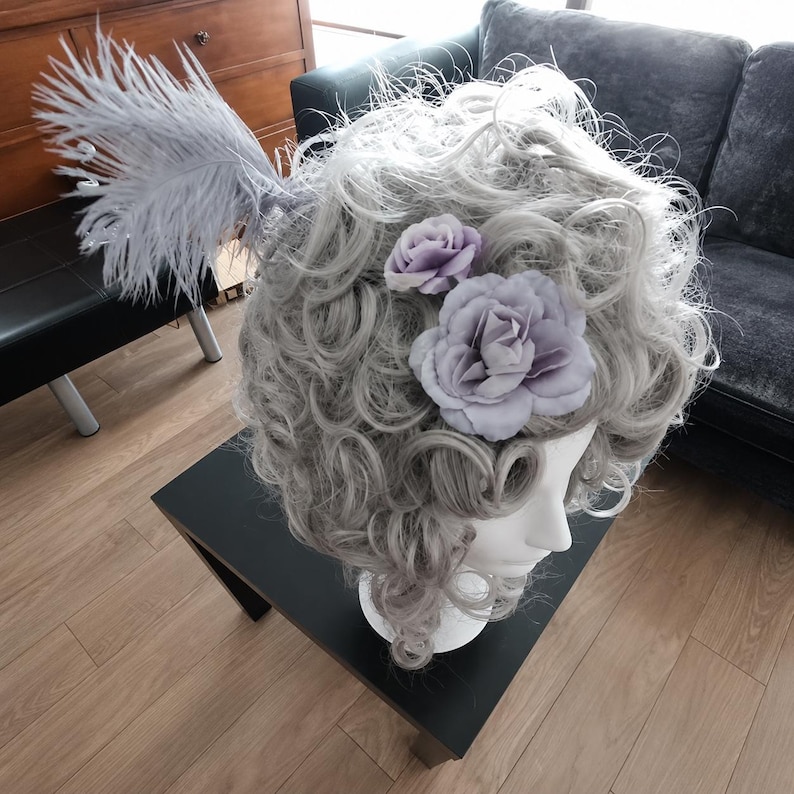 May include: A grey wig with large curls, decorated with lavender fabric flowers and a large feather plume. The wig is displayed on a white mannequin head, highlighting its elaborate style and design.