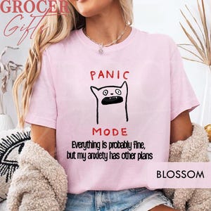 May include: A light pink short-sleeved t-shirt featuring a black line drawing of a cat with an open mouth. Above the cat is the red text PANIC and below is the red text MODE. Underneath, black text reads Everything is probably fine, but my anxiety has other plans.