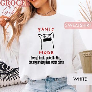 May include: A white long-sleeve sweatshirt featuring a black line drawing of a cat with an open mouth. Above the cat is the red text "PANIC MODE" and below it is the black text "Everything is probably fine, but my anxiety has other plans."