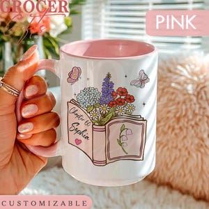 May include: A white ceramic mug with a pink interior featuring a graphic of an open book with a bouquet of colorful flowers, butterflies, and the text Chapter 60 Sophia. The mug has a pink handle and a pink rim, with a small pink heart icon.
