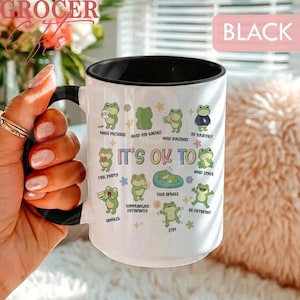 May include: White ceramic mug with a black interior and handle. The mug features a design with green cartoon frogs and the text "IT'S OK TO" with various phrases. The word "BLACK" is in a pink rectangle.