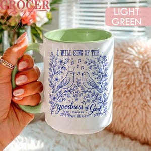 May include: A white ceramic mug with a light green interior and handle. The exterior features a blue floral design with two birds and the text: I WILL SING OF THE goodness of God PSALM 89:1. The mug has a smooth finish and a cylindrical shape.