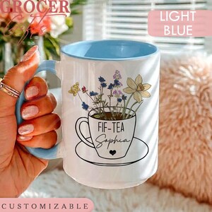 May include: A white ceramic mug with a light blue interior and handle. The mug features a graphic of a teacup filled with wildflowers and the text FIF-TEA Sophia printed in black. The design includes yellow, purple, and blue floral accents.