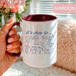 May include: White ceramic mug with a maroon interior and handle. The mug features the text "it's okay to" with various affirmations and frog illustrations in blue. The word "MAROON" is in a pink rectangle.