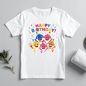 Peut inclure: T-shirt blanc avec un motif color&eacute; de requins de dessins anim&eacute;s portant des chapeaux de f&ecirc;te et le texte "HAPPY BIRTHDAY!". Le motif comprend des confettis et des serpentins, parfait pour un anniversaire.