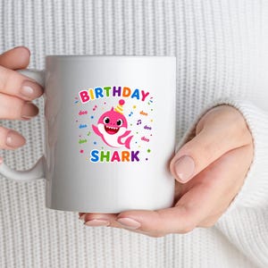 Peut inclure: Mug en c&eacute;ramique blanche avec un graphisme color&eacute; d'un requin de dessin anim&eacute; portant un chapeau de f&ecirc;te. Le texte "BIRTHDAY SHARK" est affich&eacute; au-dessus et en dessous de l'illustration du requin. Le design comprend des notes de musique et des confettis.