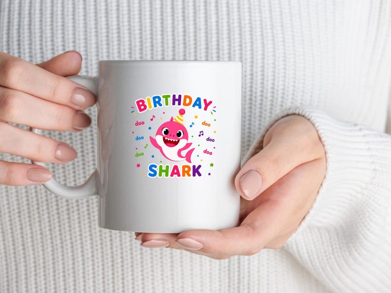Peut inclure: Mug en c&eacute;ramique blanche avec un graphisme color&eacute; d'un requin de dessin anim&eacute; portant un chapeau de f&ecirc;te. Le texte "BIRTHDAY SHARK" est affich&eacute; au-dessus et en dessous de l'illustration du requin. Le design comprend des notes de musique et des confettis.