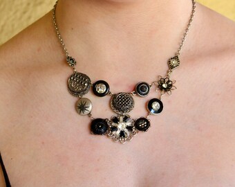 May include: A silver-toned statement necklace featuring a variety of vintage buttons and decorative elements. The necklace includes round, starburst, and floral designs in black, silver, and clear accents. The necklace is a unique piece of jewelry.