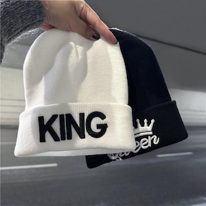 Puede incluir: Un gorro de punto blanco con la palabra KING bordada en letras negras sobre el dobladillo, junto a un gorro de punto negro con la palabra Queen bordada en cursiva blanca bajo un pequeño icono de corona en el dobladillo doblado.