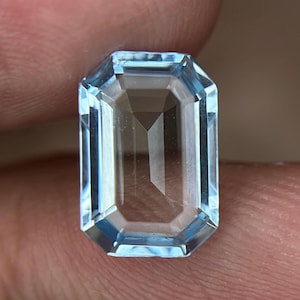 以下が含まれることがあります： 透明感のあるライトブルーの長方形のエメラルドカットの宝石。石には複数のファセットが施されており、幾何学的な反射と全体的に透明で結晶のような外観を生み出し、表面は磨き上げられています。