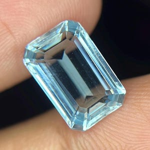 以下が含まれることがあります： エメラルドカットが施された長方形の宝石で、半透明の淡い青色をしています。石の表面には鋭く鮮明なファセットが刻まれており、光と影の幾何学的な模様を作り出し、その透明で結晶のような構造を際立たせています。