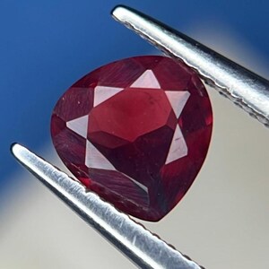 May include: A deep red, pear-shaped faceted gemstone held by silver-toned metal tweezers. The stone features a clear, symmetrical cut with multiple triangular and trapezoidal facets that reflect light across its polished, translucent surface.
