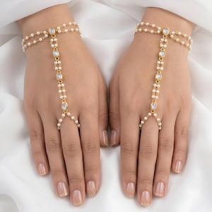 Puede incluir: Una pulsera de mano dorada con delicadas perlas blancas y piedras ovaladas iridiscentes. La pieza de joyería consta de una pulsera de muñeca de doble cadena conectada por una cadena central de perlas y piedras que llega a un anillo.