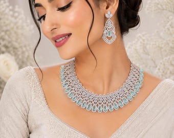 Op de afbeelding: Een zilverkleurige ketting en bijpassende oorbellen met ingewikkeld filigraanwerk en ingelegde lichtblauwe, peervormige stenen. De ketting heeft een breed, gebogen ontwerp en de oorbellen hebben een hangende druppelvorm.