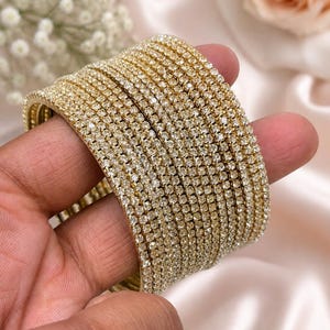 Peut inclure: Un ensemble de plusieurs bracelets joncs dorés, chacun orné d'une rangée unique de strass transparents et scintillants incrustés sur la surface extérieure. Ces bracelets fins, circulaires et métalliques sont conçus pour être portés ensemble.