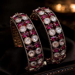 May include: Two gold-toned bangles featuring a repeating pattern of round clear stones and rectangular deep red stones. The metal framework has a textured, beaded edge design, and each bangle includes a small hinged clasp mechanism for closure.