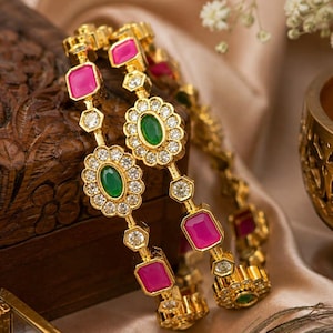 May include: Two gold-toned bangles featuring alternating rectangular fuchsia stones and oval green stones surrounded by small clear white crystals. The metal links connect each stone setting in a continuous circular design for the wrist.