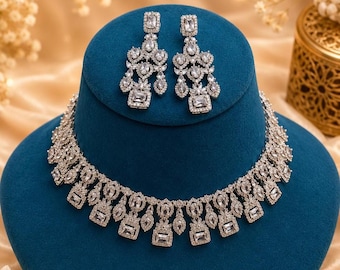 May include: A silver-toned jewelry set consisting of a necklace and a pair of matching drop earrings. The pieces feature clear, rectangular and teardrop-shaped stones arranged in a repeating pattern, set in a metallic frame with a polished finish.