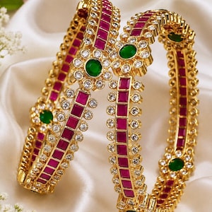 May include: Two gold bangles featuring a central row of square-cut magenta stones flanked by small clear round stones. The design includes circular floral motifs with a central round green stone surrounded by clear stones at regular intervals.