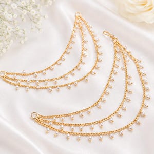 Op de afbeelding: Twee goudkleurige metalen kettingaccessoires met meerdere gedrapeerde lagen. Elke ketting is versierd met kleine, gelijkmatig verdeelde witte kralen. De stukken hebben ronde metalen ringen aan de uiteinden voor bevestiging.