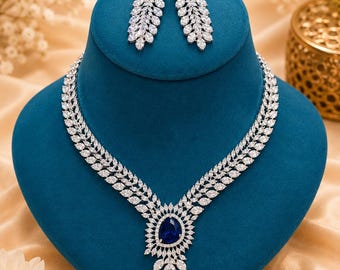 May include: A jewelry set consisting of a necklace and matching earrings. The necklace features a central pear-shaped blue stone surrounded by clear marquise-cut stones in a leaf-like pattern. The earrings mirror this design with a blue stone top.
