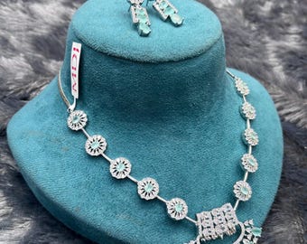 May include: A silver-toned necklace and earring set featuring light blue oval-cut stones surrounded by small clear crystals. The necklace has a central semi-circular pendant with dangling stones, and the earrings are matching stud-style pieces.