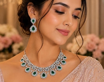 Peut inclure: Un collier et des boucles d'oreilles assorties en métal argenté, ornés de pierres vertes en forme de poire entourées de petites pierres claires. Le collier présente un motif festonné avec plusieurs pierres vertes pendantes.