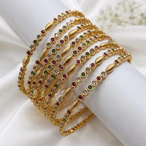 May include: A set of gold-toned bangles featuring a repeating pattern of small, round, bezel-set stones in red, green, and clear white, interspersed with textured, ribbed gold-toned metal segments along the circumference of each circular bracelet.