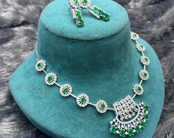 May include: A silver-toned necklace and matching earring set featuring oval-cut green stones surrounded by small clear crystals. The necklace has a central fan-shaped pendant with a grid of clear crystals and a row of green stones along the bottom edge.