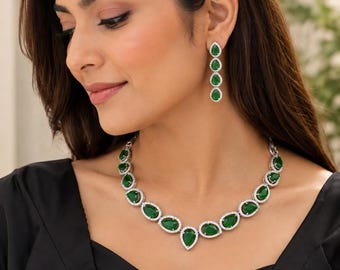 Puede incluir: Un conjunto de collar y pendientes colgantes con piedras verdes en forma de pera rodeadas de pequeñas piedras blancas transparentes en un engaste de metal plateado. El collar consta de una serie de estas piedras unidas entre sí.