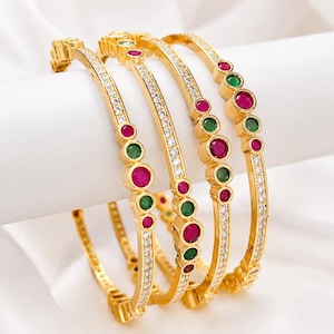 Peut inclure: Un ensemble de quatre bracelets rigides dorés ornés d'une rangée centrale de petites pierres claires. Chaque bracelet est agrémenté de pierres rondes rouges et vertes alternées, serties dans des chatons dorés sur le bord extérieur.