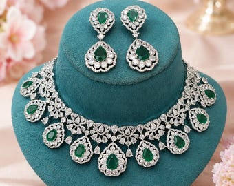 May include: A jewelry set consisting of a necklace and matching drop earrings. The pieces feature pear-shaped green stones surrounded by clear, round-cut stones set in a silver-toned metal frame, creating a floral and geometric pattern.