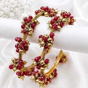 Peut inclure: Une paire de bracelets rigides dorés dotés d'une bande métallique texturée ornée de grappes de petites perles facettées rouge foncé et de minuscules sphères métalliques dorées. Le design intègre des motifs floraux de perles répétitifs.