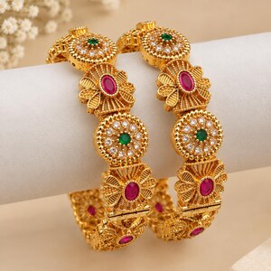May include: Two gold-toned bangles featuring an intricate filigree design. Each bangle is adorned with alternating circular motifs containing clear white stones and a central green stone, alongside floral-shaped elements set with oval pink stones.