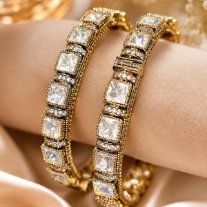 May include: Two gold-toned bangles featuring a series of square-cut clear stones set in a repeating pattern. Each bangle is accented with small, pavé-style clear stones along the edges, creating a textured, metallic finish with a bright, reflective appearance.