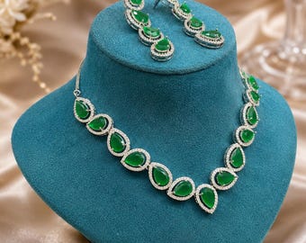 May include: A jewelry set consisting of a necklace and a pair of drop earrings. The necklace features a series of pear-shaped green stones surrounded by small clear crystals in a silver-toned setting. The earrings match with a similar design.