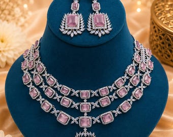 Op de afbeelding: Een driedubbele ketting en bijpassende oorbellen met rechthoekige lichtroze stenen omringd door heldere witte kristallen in een zilverkleurige metalen zetting, inclusief een centrale rechthoekige hanger aan de ketting.