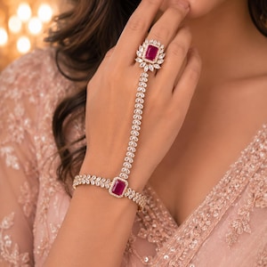 Könnte beinhalten: Ein Handrückenarmband mit zwei rechteckigen, dunkelrosa Steinen in goldfarbener Fassung. Ring und Armband sind durch eine Kette aus klaren, marquise-geschliffenen Steinen verbunden, wobei die rosa Edelsteine von weiteren klaren Steinen umrandet sind.