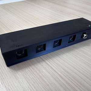 May include: A rectangular black electronic signal switcher featuring four circular ports on the front face, one silver-colored jack, and two small indicator lights in green and red. The word INPUT is embossed on the left side of the device.