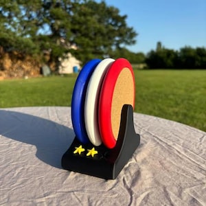 Puede incluir: Un juego de tres posavasos coloridos en azul, blanco y rojo, exhibidos en un soporte negro con dos estrellas amarillas. Los posavasos tienen un centro de corcho y están diseñados para proteger las superficies.