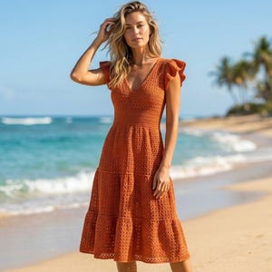 May include: A rust-colored midi dress crafted from crochet fabric, featuring a V-neckline, short flutter sleeves, and a tiered skirt design. The garment has a fitted bodice that transitions into a flared, textured skirt with a consistent knit pattern.
