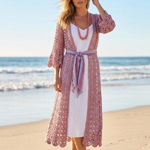 May include: A long, open-front mauve crochet cardigan featuring intricate openwork patterns, scalloped edges, and three-quarter length sleeves, worn over a simple white dress with a light purple fabric sash tied at the waist and a beaded necklace.