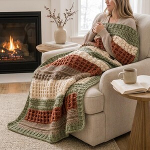 May include: A crocheted throw blanket featuring horizontal stripes in sage green, cream, and burnt orange. The blanket displays a textured waffle stitch pattern interspersed with rows of bobble stitches, creating a thick, cozy, and layered appearance.