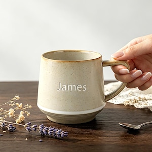 Op de afbeelding: Een gespikkelde beige keramische mok met een taps toelopende voet en een handvat, voorzien van de naam James in witte letters op de voorzijde. De mok heeft een gladde afwerking met een subtiele bruine rand en een eenvoudig ontwerp.