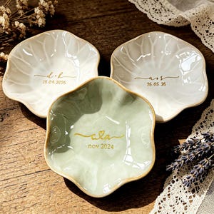 May include: Three small, scalloped-edge ceramic trinket dishes. Two are white with gold lettering reading d+k 18.04.2026 and a+s 26.05.26. The third is sage green with gold lettering reading cla nov 2024. Each dish has a glossy, wavy surface.