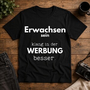 Könnte beinhalten: Ein schwarzes T-Shirt mit kurzen Ärmeln und dem weißen Text: "Erwachsen sein klang in der WERBUNG besser." Das Shirt hat einen Rundhalsausschnitt und besteht aus einfarbigem schwarzem Stoff mit kontrastreicher weißer Schrift auf der Vorderseite.
